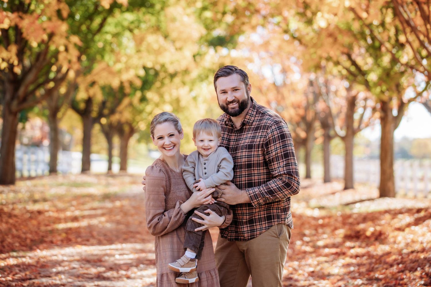 Family of three posing among autumn trees with colorful fall foliage, parents holding smiling baby outdoors.