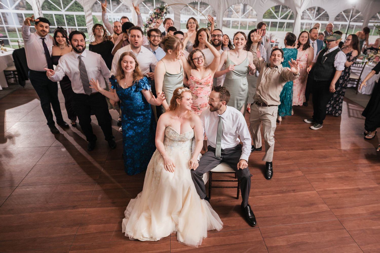 Bride and groom surrounded by cheering, dancing guests on the reception dance floor, everyone throwing their hands up together.