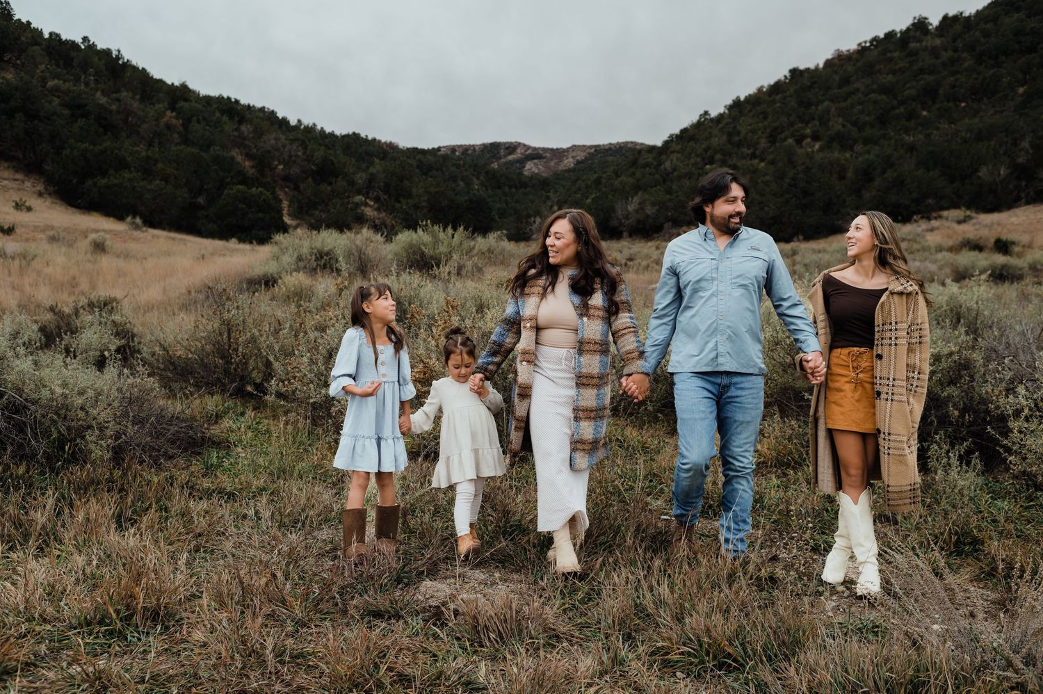 Family of five walking hand-in-hand through a scenic mountain meadow on a cloudy day.