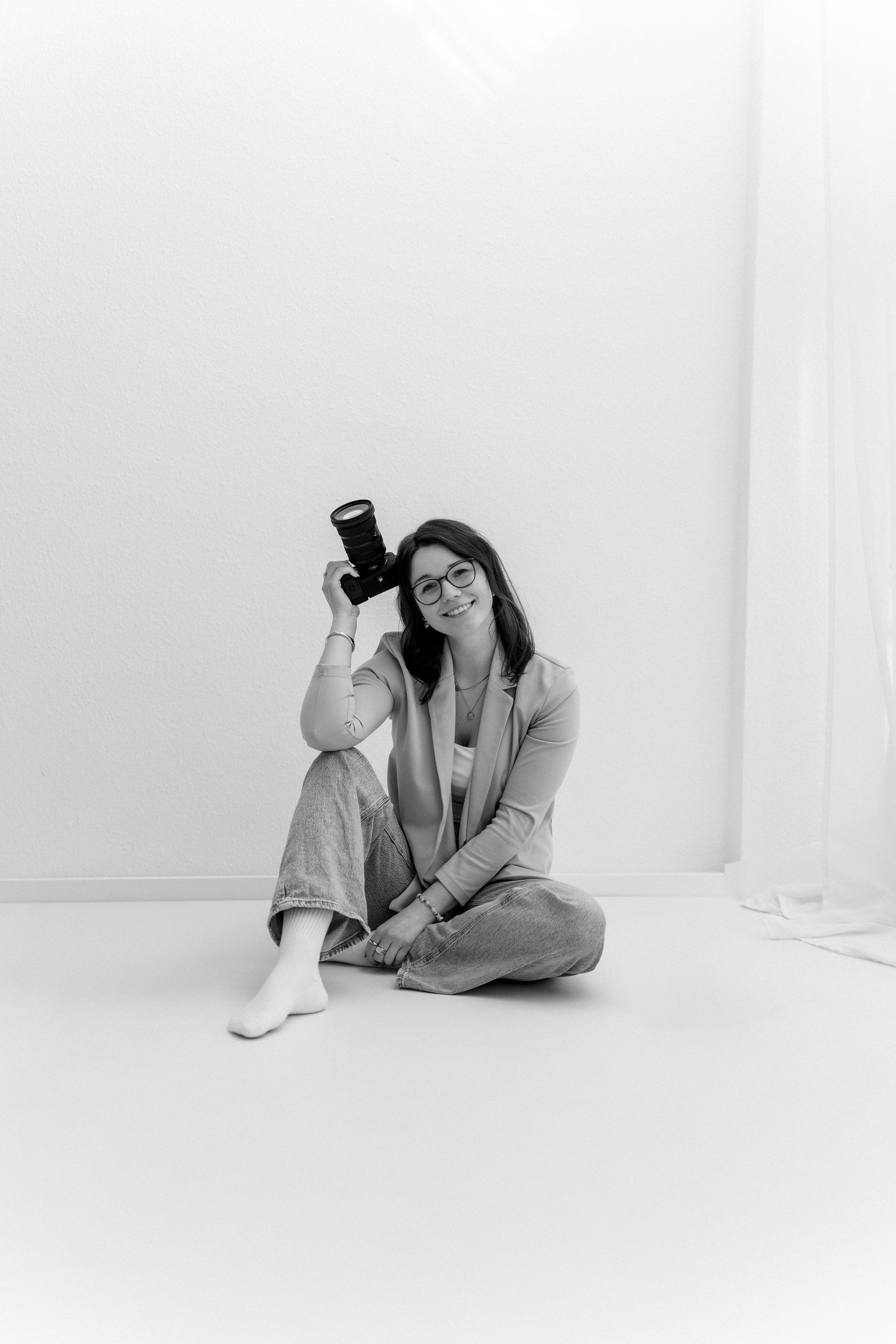 Black and white photo of a smiling woman sitting cross-legged on floor holding a camera, wearing glasses and casual outfit.