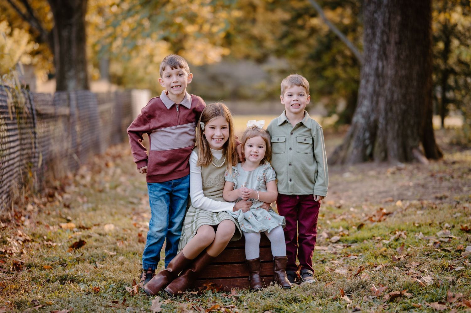 Four children posing outdoors in autumn, smiling together near a wooden crate with fall foliage backdrop.