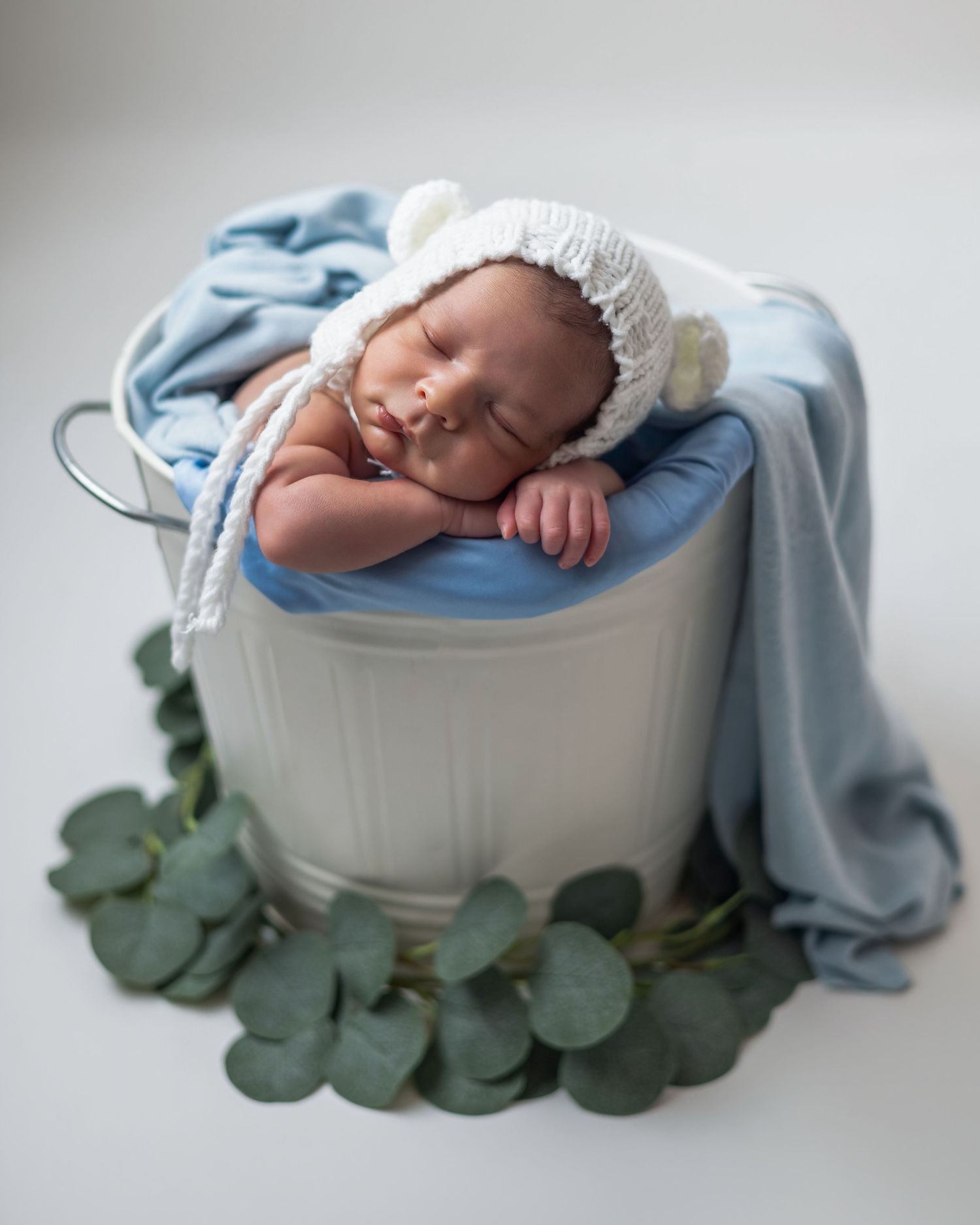 Sleeping newborn baby wearing a white knit bear bonnet, nestled in a white bucket with blue fabric and eucalyptus leaves.