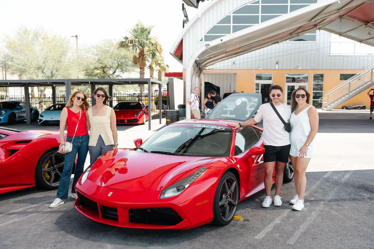 Four people posing beside a red Ferrari 488 at an exotic car dealership on a sunny day.