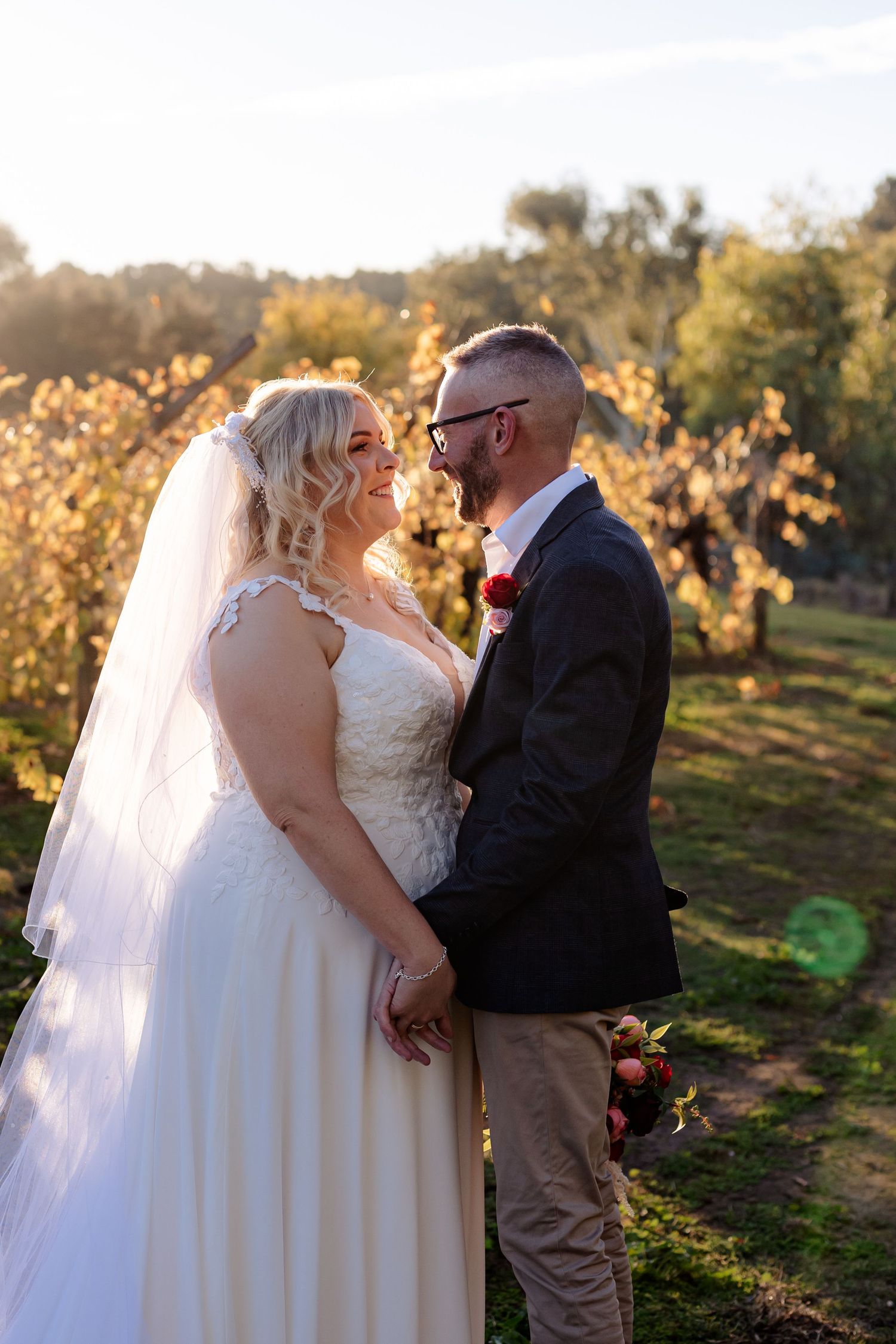Bride and groom share a romantic moment in a sunlit vineyard, bride wearing a lace gown and veil, groom in a dark blazer.