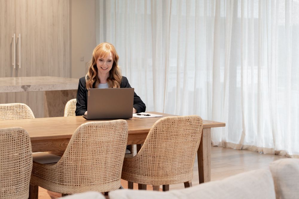 Woman with red hair working on laptop at wooden dining table in modern bright room with wicker chairs.
