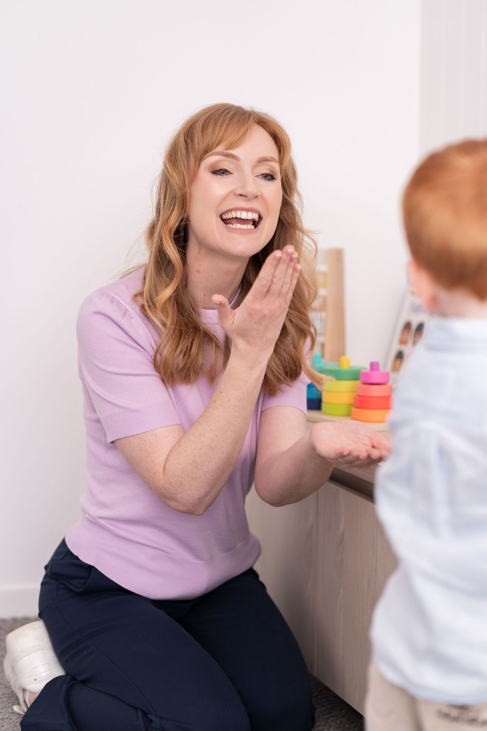 A smiling woman in a purple shirt plays patty-cake with a young redheaded toddler near colorful stacking toys.