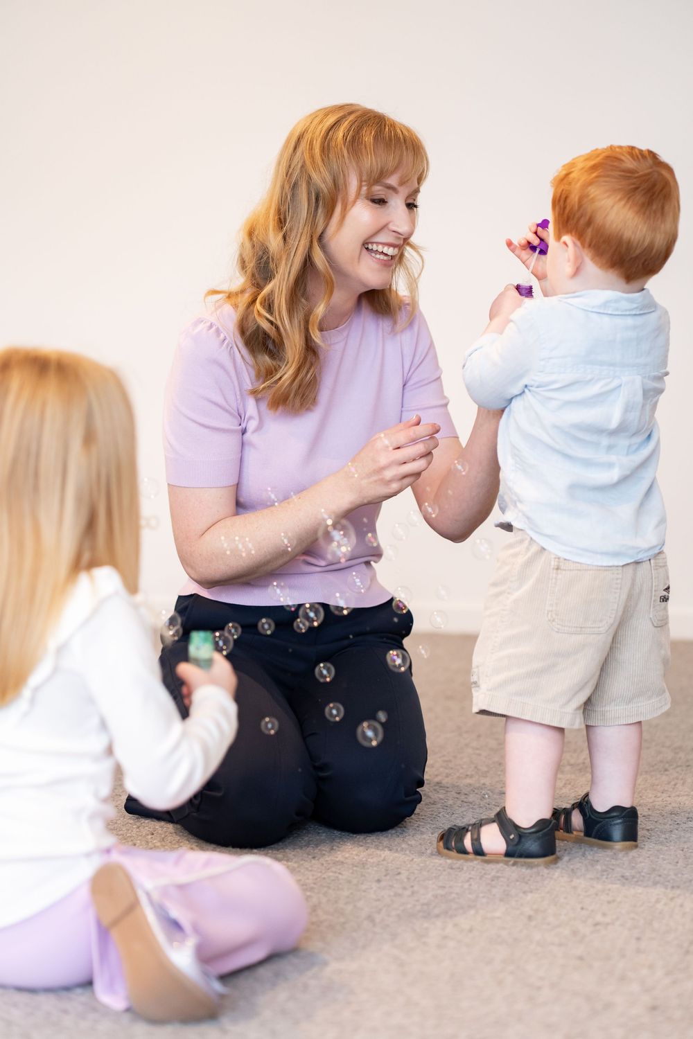 A smiling redhead woman kneels on carpet playing with two young children using bubble wands indoors.
