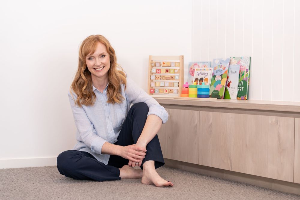 Smiling redhead woman sitting cross-legged on floor near wooden shelf with colorful children's toys and books.