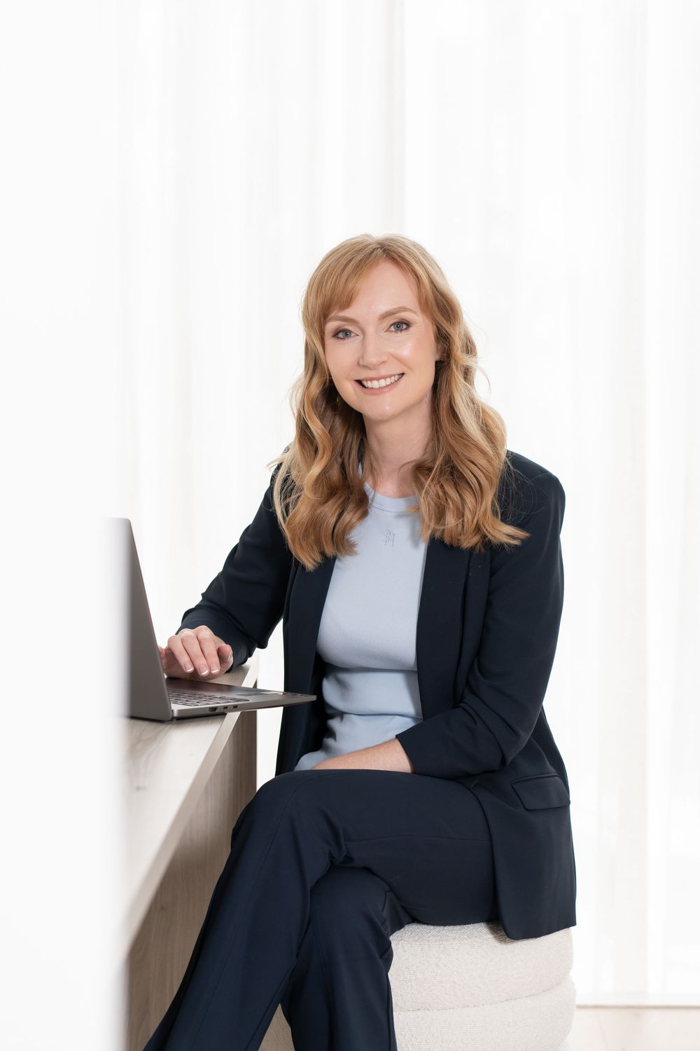 Smiling businesswoman in dark suit sitting with laptop near white curtains in bright office setting.