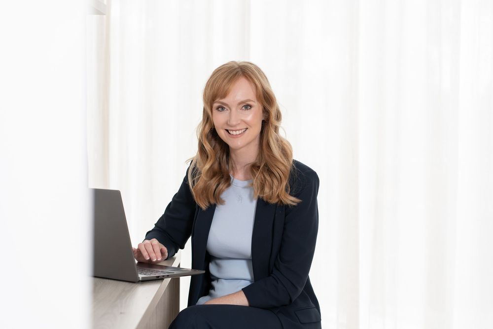 Professional businesswoman in navy blazer smiling at laptop in bright modern office setting.