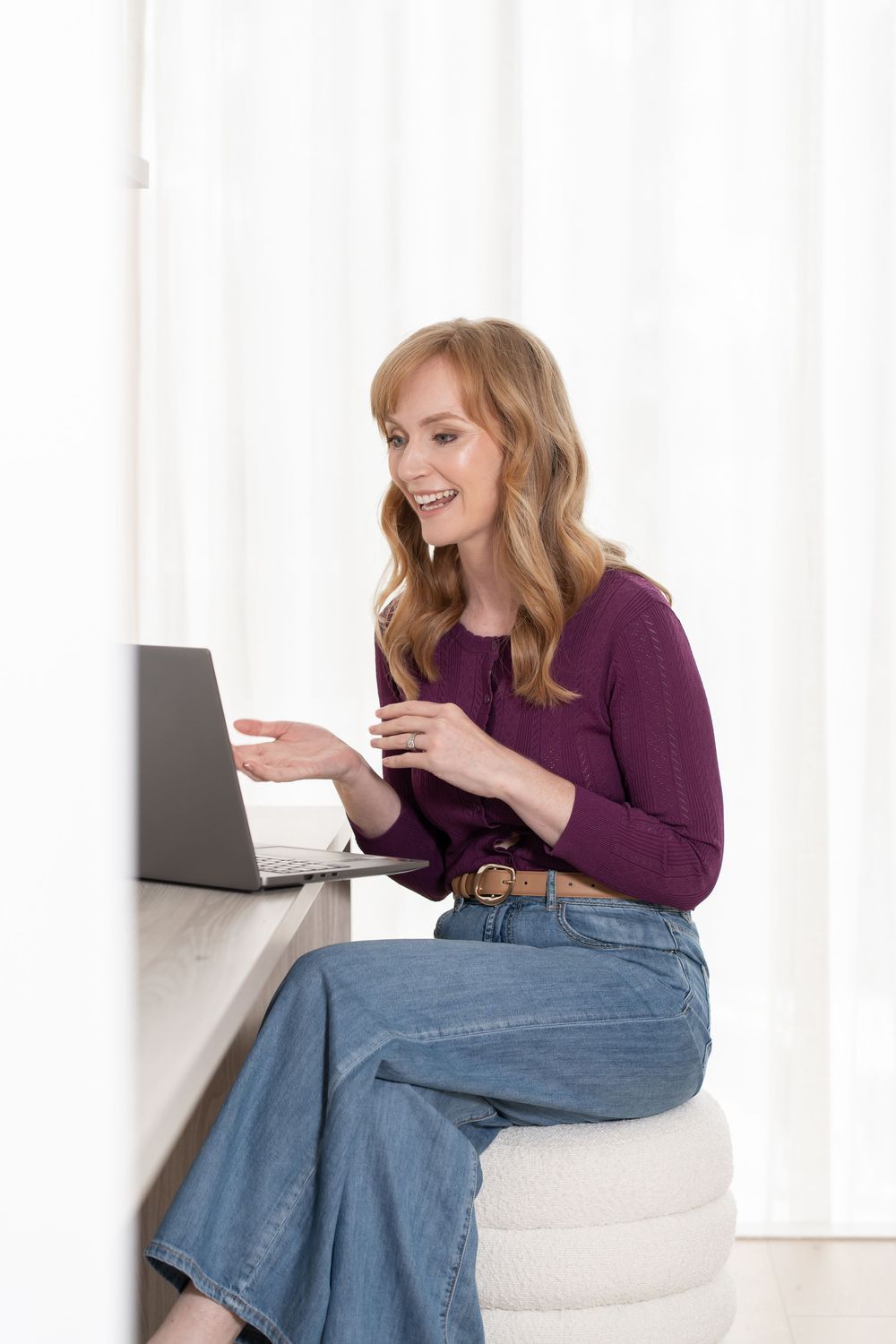Smiling woman in purple top and jeans sitting on ottoman using laptop computer at home desk.