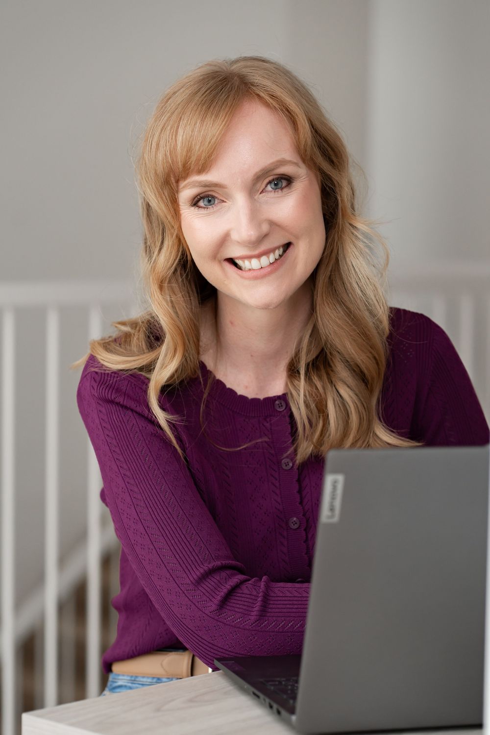 Smiling blonde woman in purple sweater working on a Lenovo laptop at a desk in a bright home office.
