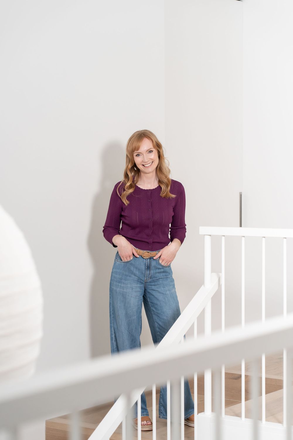 Smiling young woman with wavy blonde hair wearing a purple sweater and jeans, standing on white staircase indoors.