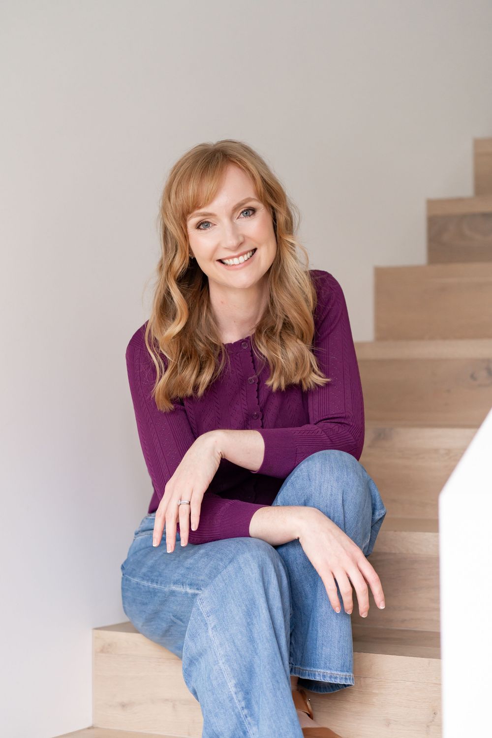 Happy redhead woman in purple top and jeans sitting on wooden stairs smiling at camera.