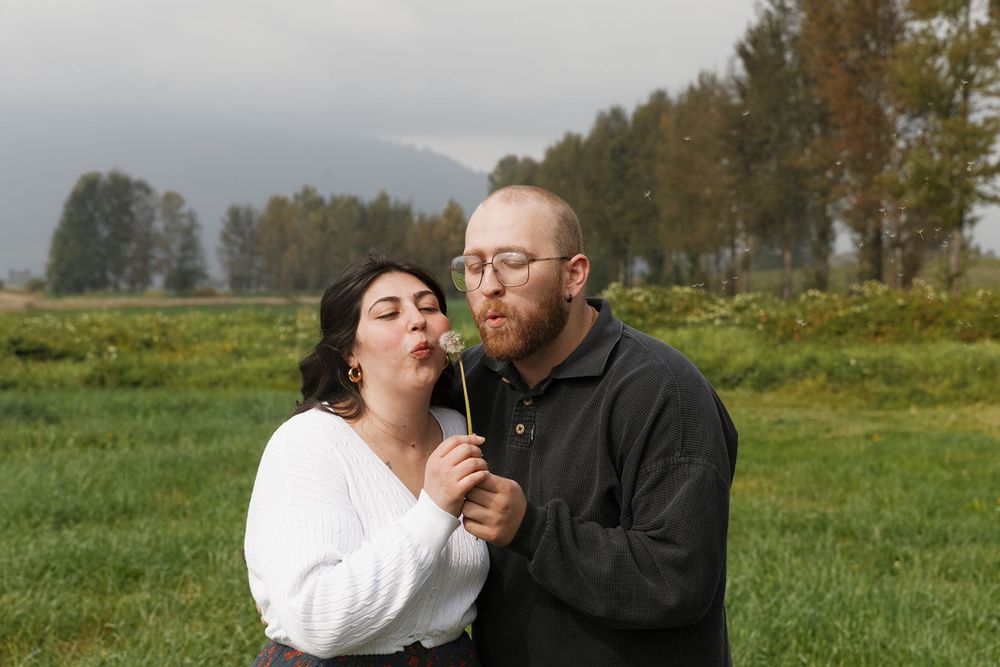 Couple blowing a dandelion together in a green field with misty mountains in the background.