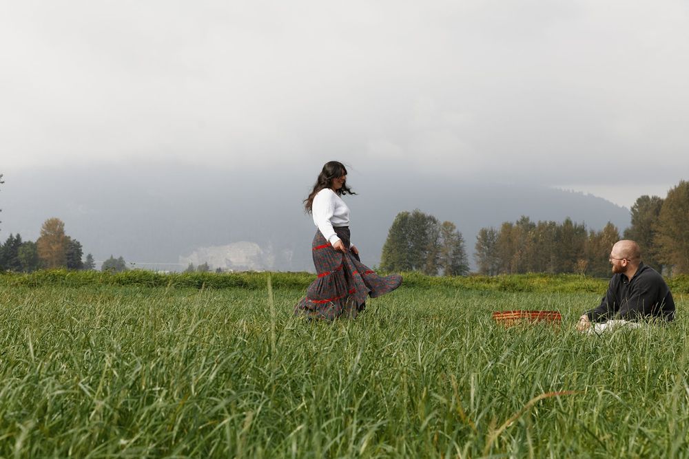 A woman in a flowing skirt walks through a lush green field while an elderly man sits nearby under cloudy skies.