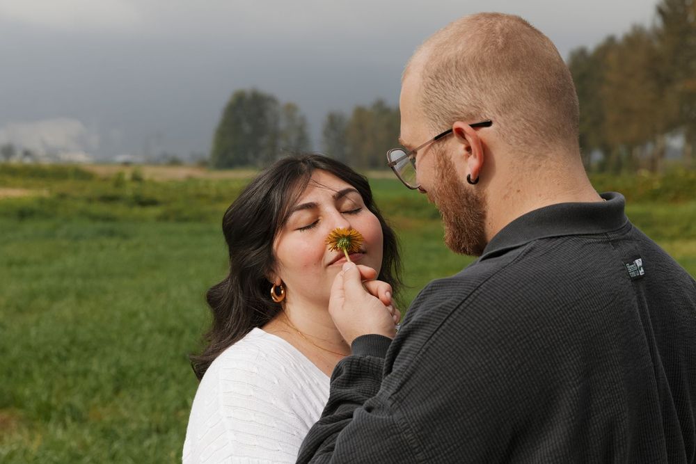 A man tenderly holds a small flower to his smiling partner's nose in a green field.