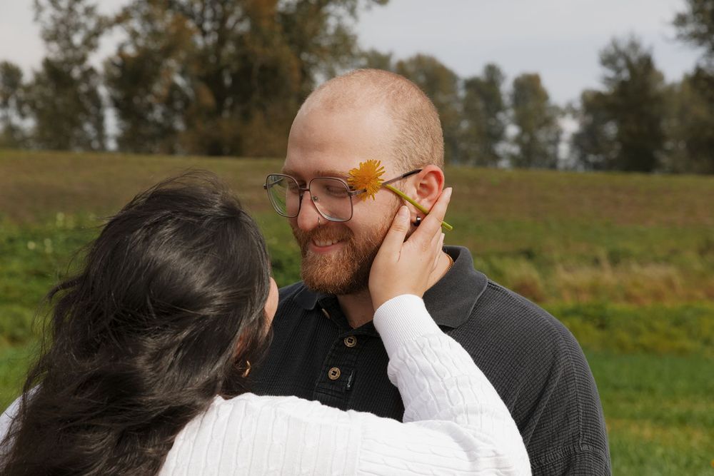 A bearded man smiles as a woman places a yellow flower on his cheek outdoors in a green field.