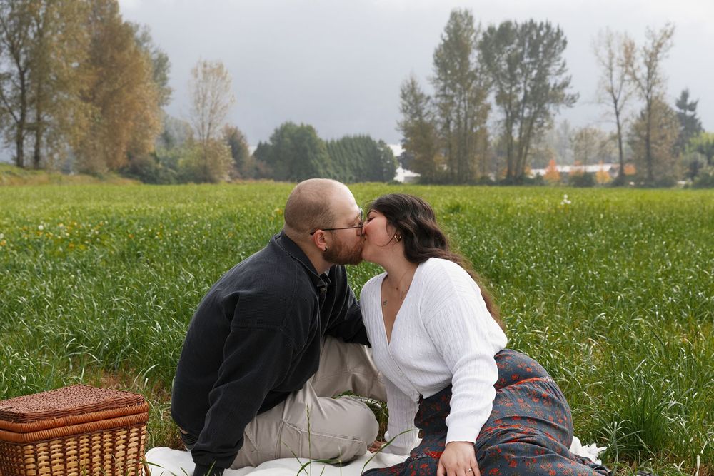 Couple sharing a romantic kiss during a picnic in a lush green field with trees in the background.