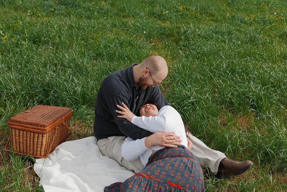 Couple sharing a tender embrace on a white blanket during a picnic in a lush green field with a wicker basket nearby.