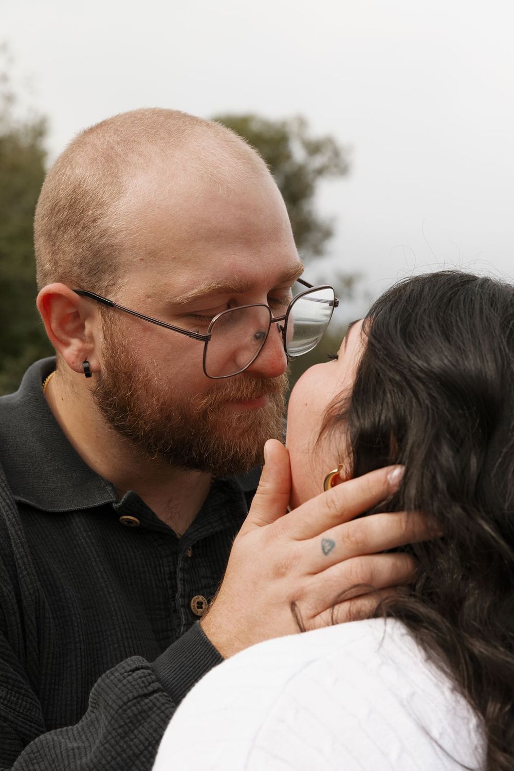 Bearded man with glasses tenderly holds woman's face as they share a romantic moment outdoors.