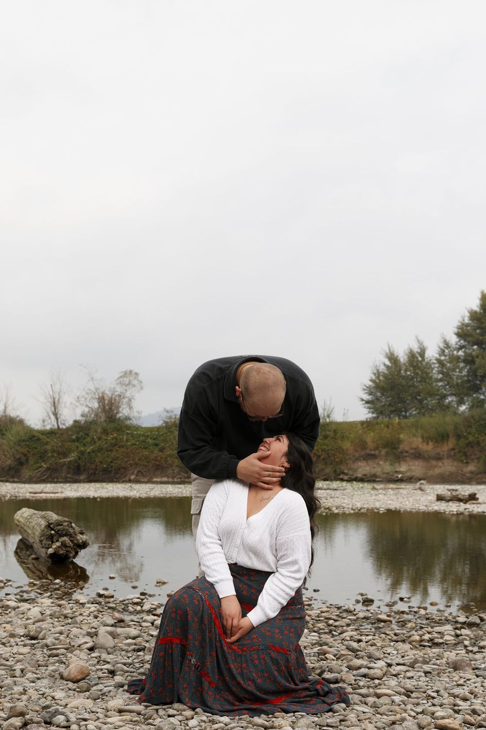 A bald man tenderly kisses a seated woman near a rocky riverbank on an overcast day.