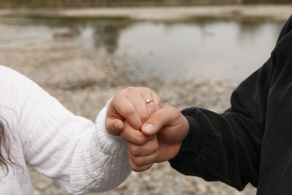 Couple holding hands outdoors near water, woman wearing gold engagement ring, romantic close-up shot.