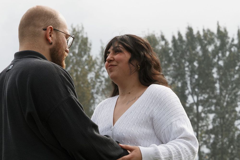 A couple shares a tender moment outdoors, the woman in a white sweater smiling warmly at her partner in a black jacket.