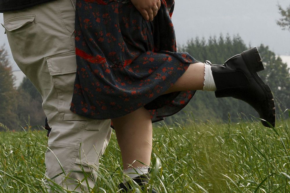 A couple embraces in a lush green field, the woman lifted slightly wearing a floral dress and black boots.