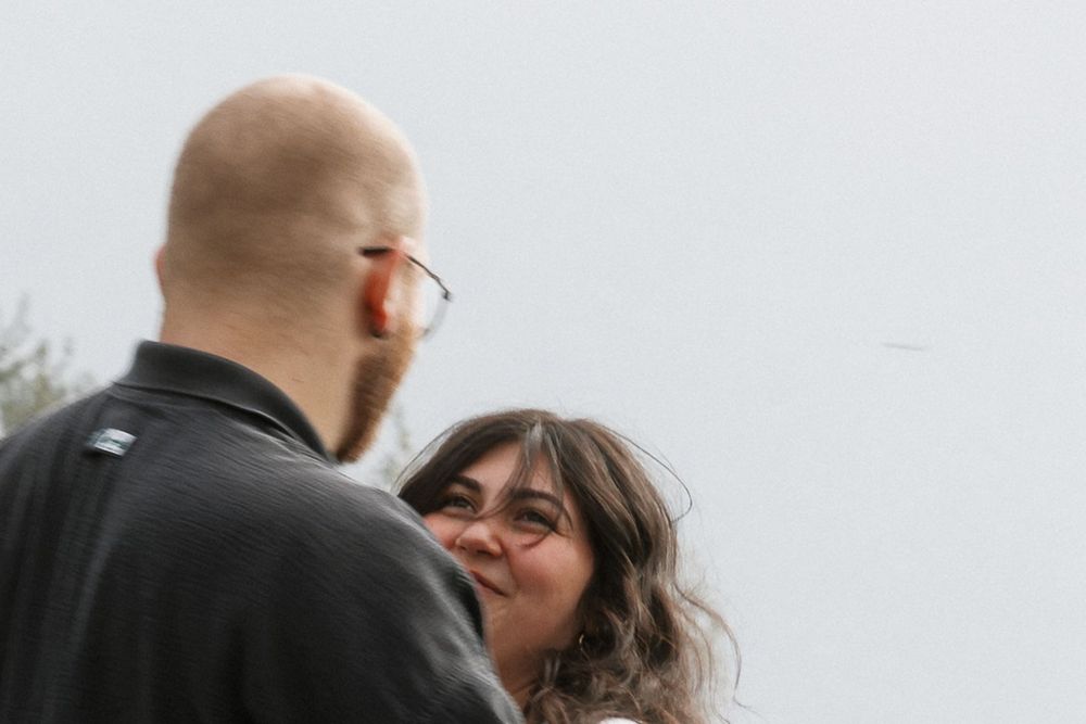 A smiling woman with dark hair gazes up at a bald bearded man wearing glasses during an outdoor couples session.