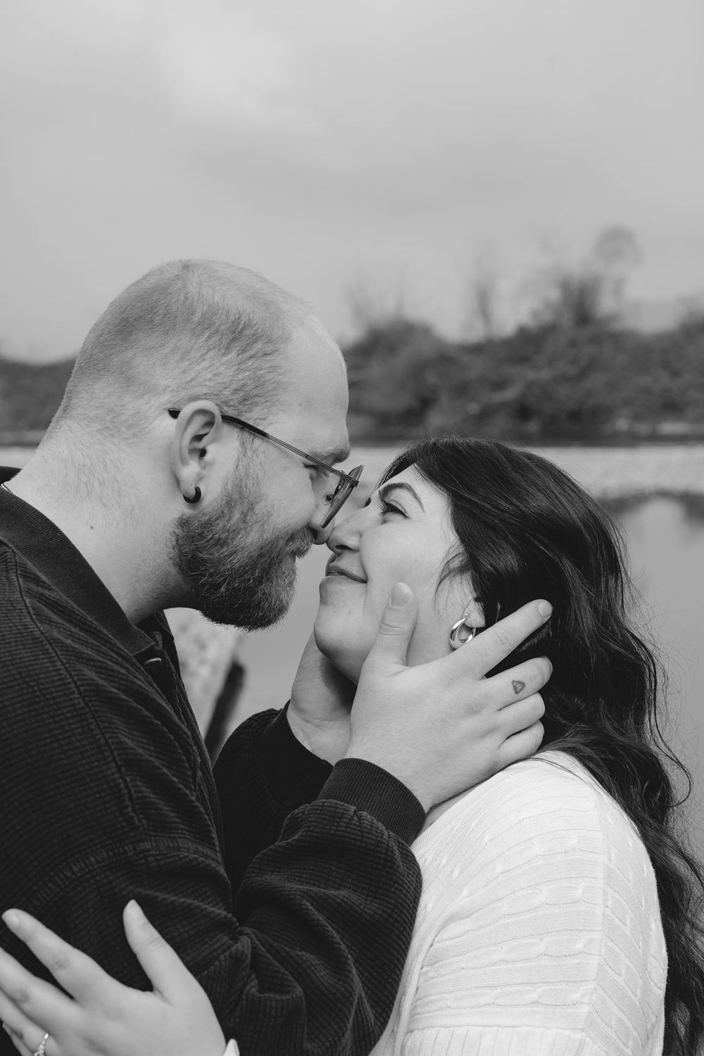 A black and white photo of a couple sharing an intimate almost-kiss near a river.