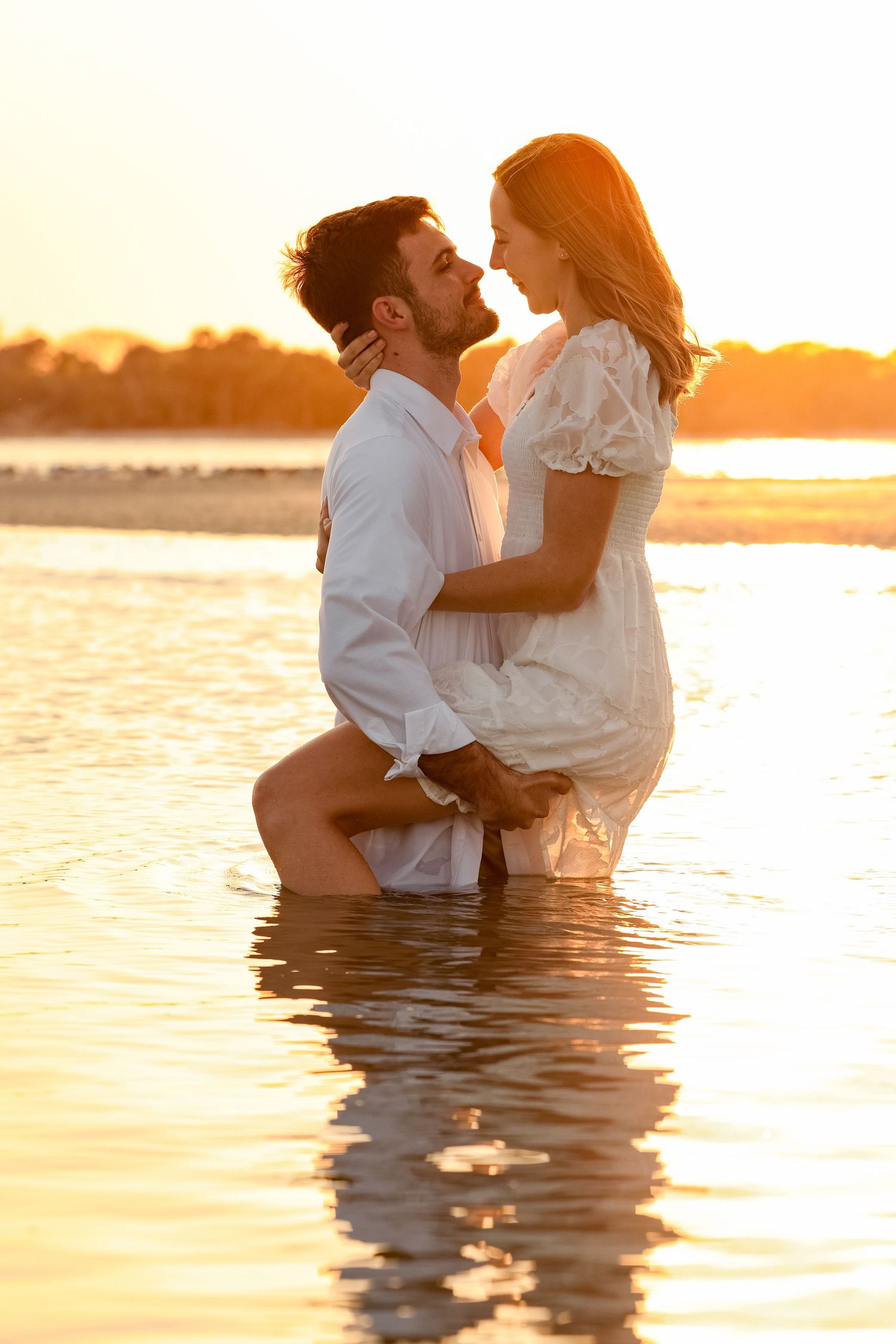 Romantic couple embracing in golden sunset water, woman in white dress held by man in white shirt.