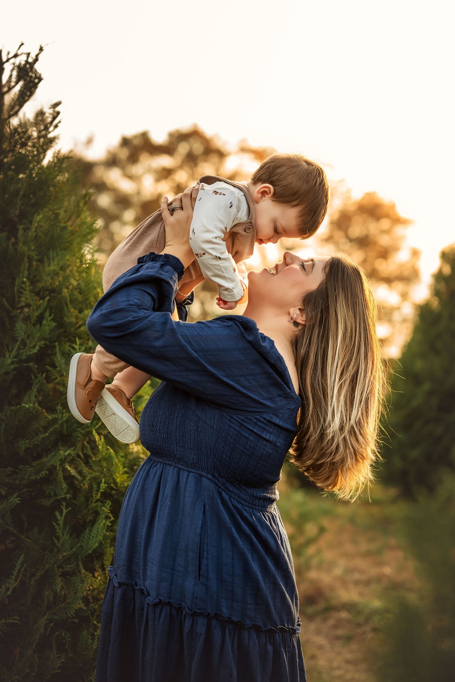 A mother in a blue dress lifts her toddler at golden hour in a lush green tree farm setting.