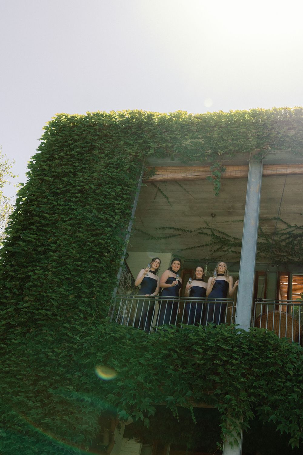 Four bridesmaids in navy dresses stand on an ivy-covered balcony in soft sunlight.
