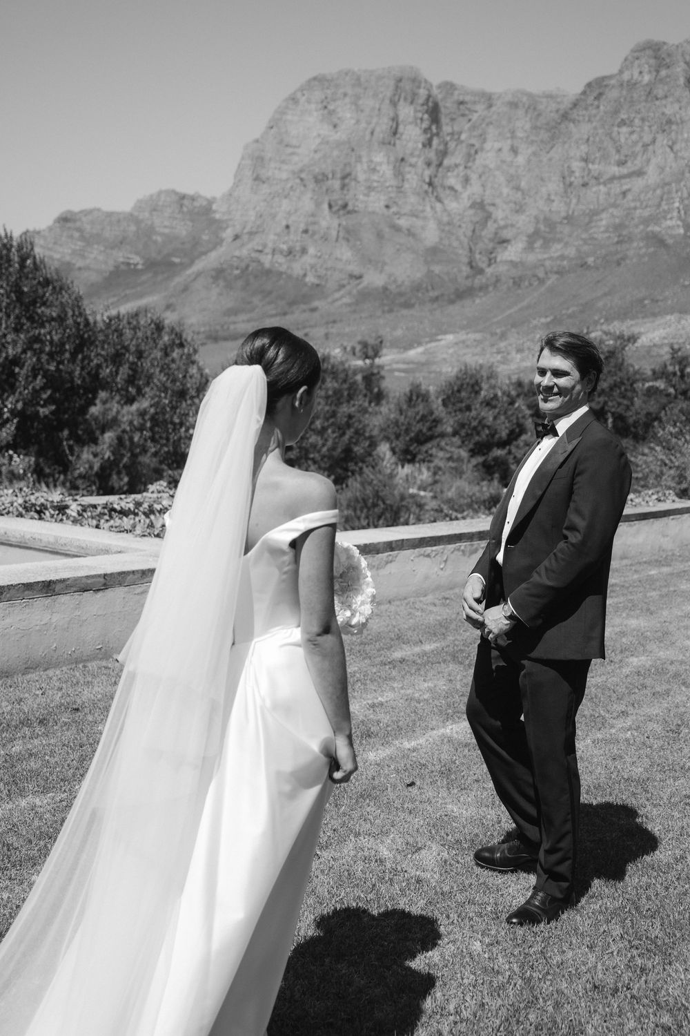 Black and white photo of a smiling groom turning to face his bride in a wedding gown with veil, mountains in background.
