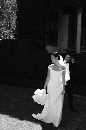 Black and white photo of bride in off-shoulder gown holding white bouquet with groom in tuxedo in garden.