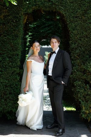 Bride in white off-shoulder gown and groom in tuxedo posing under green hedge archway outdoors.
