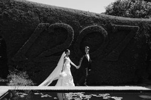 Black and white wedding photo of bride and groom holding hands in front of a hedge sculpture reading 227.