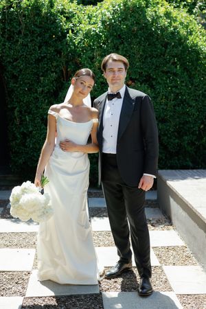 Bride in white off-shoulder gown holding bouquet stands with groom in black tuxedo outdoors.
