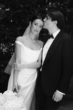 Black and white wedding photo of bride in off-shoulder gown with veil, groom in tuxedo kissing her cheek.