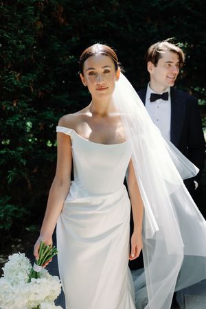 Bride in off-shoulder white gown and veil holds white bouquet, groom in navy tuxedo behind her outdoors.
