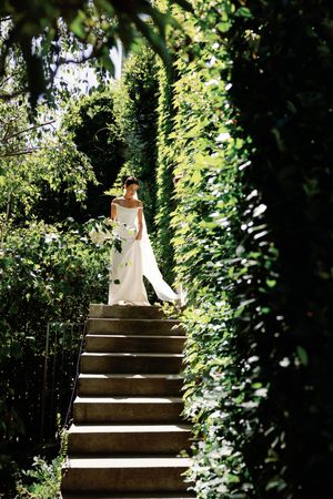 Bride in white gown descending stone steps surrounded by lush green hedges in a garden setting.