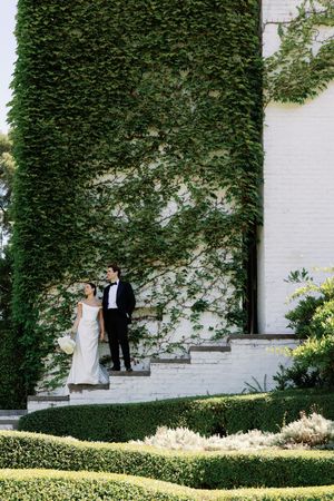 Bride and groom descend ivy-covered white brick steps surrounded by manicured hedges in elegant wedding attire.