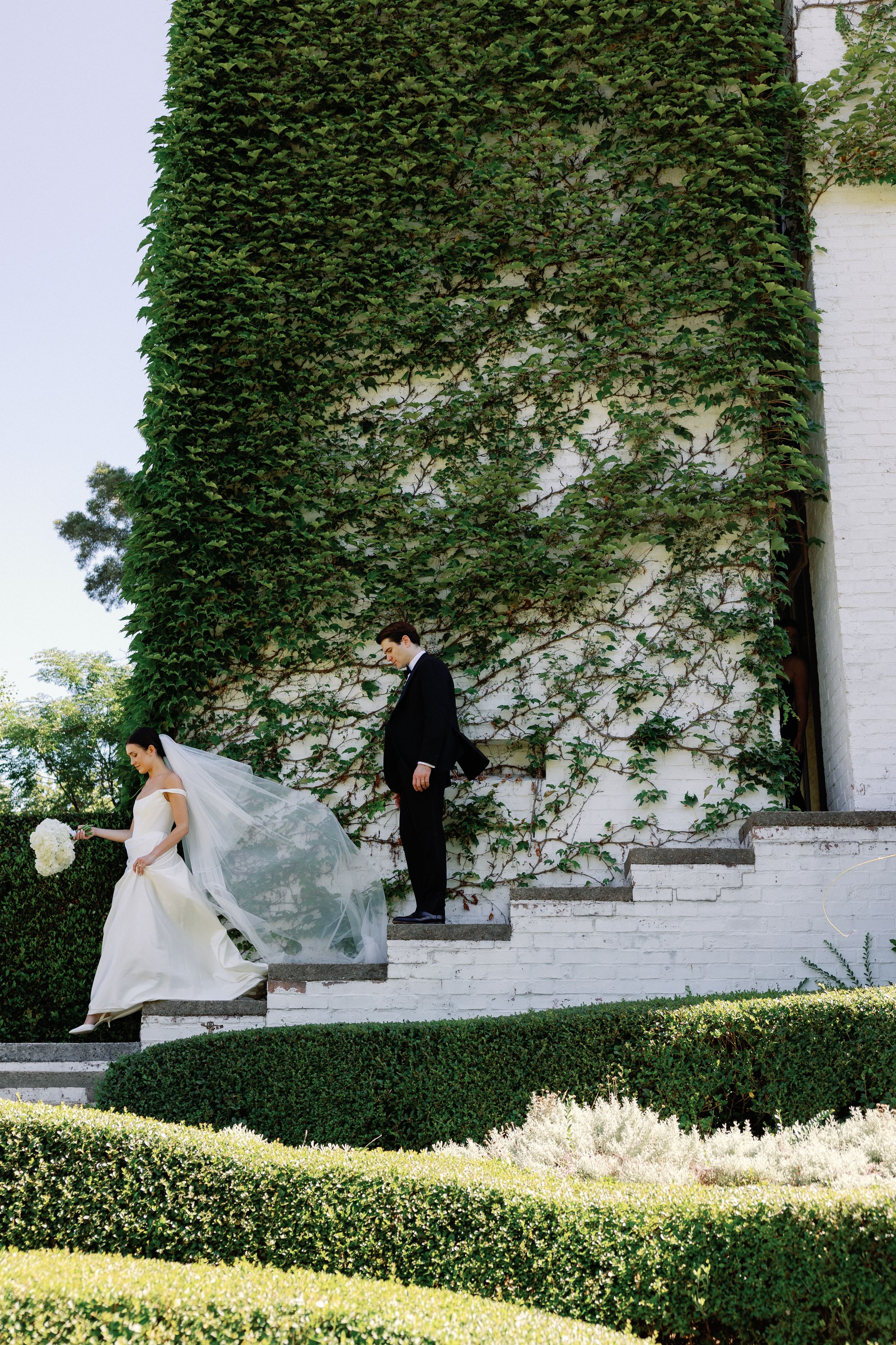 Bride and groom descend ivy-covered white brick steps in a lush garden, her veil flowing in the breeze.