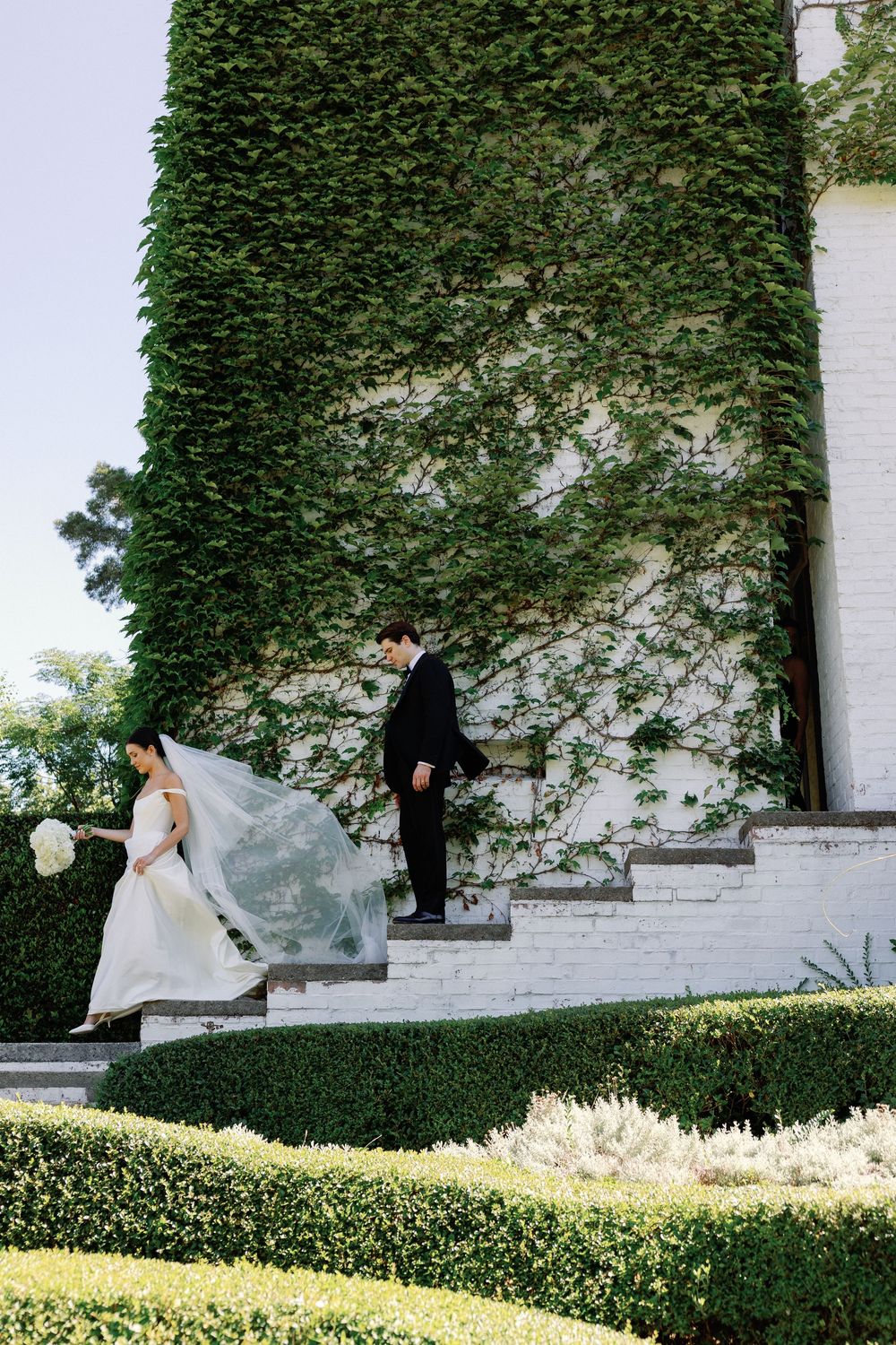 Bride and groom descend ivy-covered white brick steps in a lush garden, her veil flowing in the breeze.