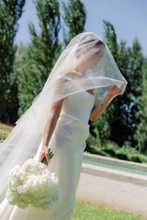 Bride in white gown lifts flowing veil outdoors, holding white hydrangea bouquet near garden fountain.