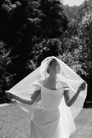 Black and white photo of a bride in a white gown lifting her veil outdoors with lush green trees in the background.