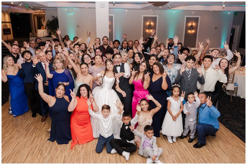 Large wedding group photo with bride and groom surrounded by guests in formal attire, celebrating in a decorated banquet hall.