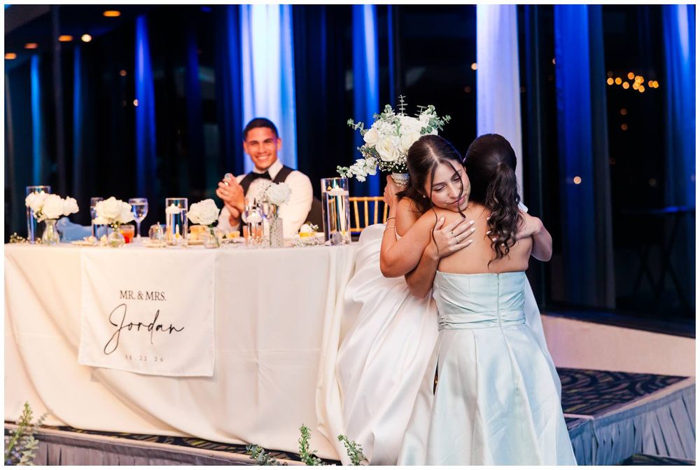 Bride shares a warm hug with a guest at wedding reception near a sweetheart table with floral arrangements.