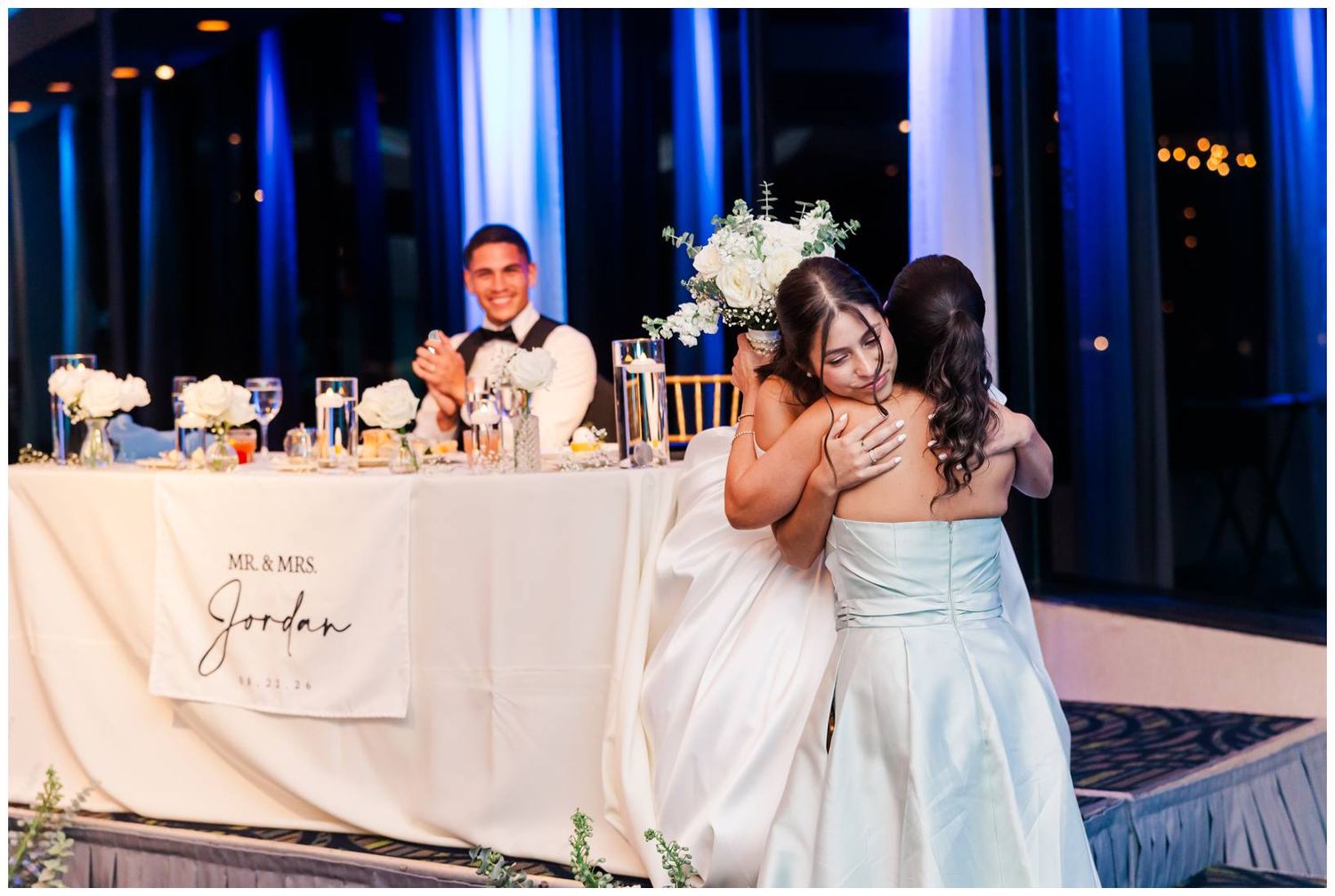 Bride shares a warm hug with a guest at wedding reception near a sweetheart table with floral arrangements.